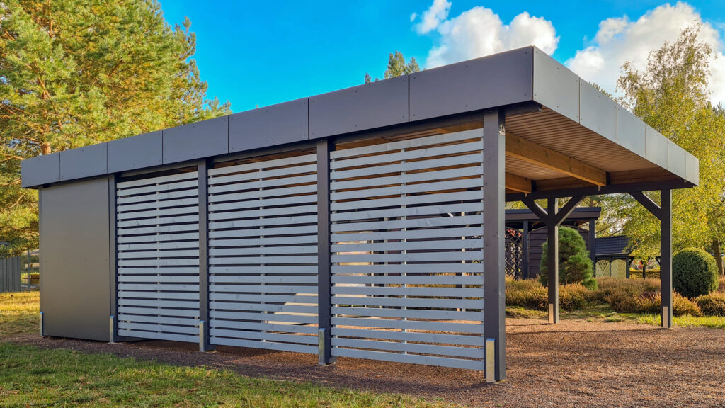 Moderner Carport mit grauen Lamellenwänden und Flachdach, umgeben von Bäumen und Sträuchern