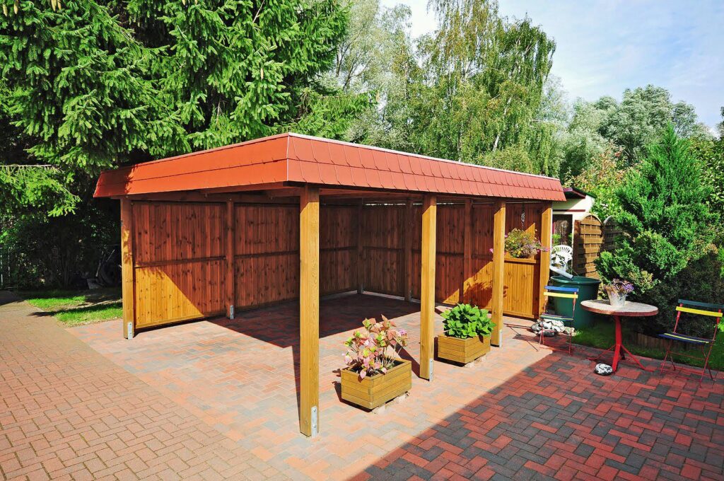 Attraktiver Flachdach-Einzelcarport aus Holz mit roter SchindelblendeEinzelcarport aus Holz mit rotem Dach, umgeben von Bäumen und bepflanzten Holzkästen auf gepflastertem Boden