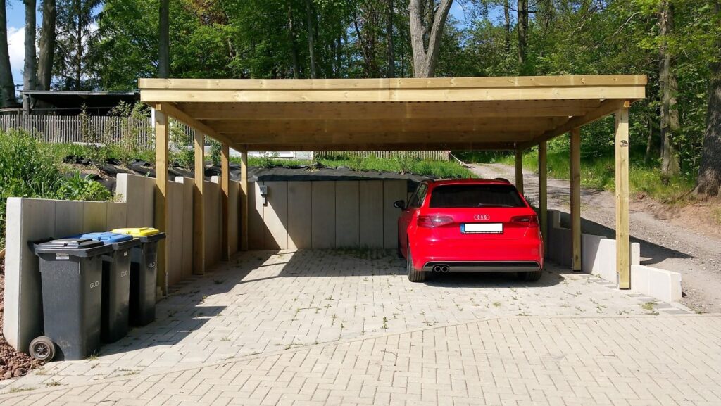 Moderner Flachdach-Doppelcarport freitragend aus HolzDoppelcarport aus Holz mit Flachdach darunter parkt ein roter PKW, umgeben von Bäumen