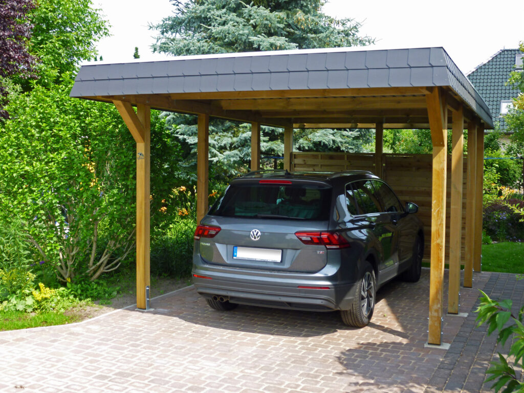 Holzcarport mit grauem Flachdach, darin geparkt ein grauer SUV, umgeben von grünem Garten