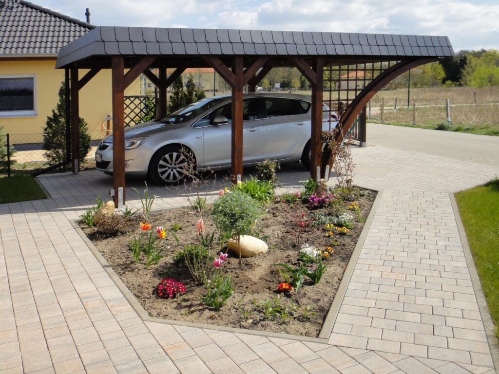 Rundbogen Einzel-Carport mit dunkler Schindelblende schützt ein silbernes Auto auf der gepflasterten Einfahrt