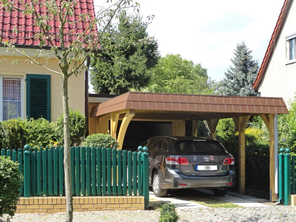 Rundbogen Carport aus Holz mit brauner Schindelblende und parkendem Auto darunter