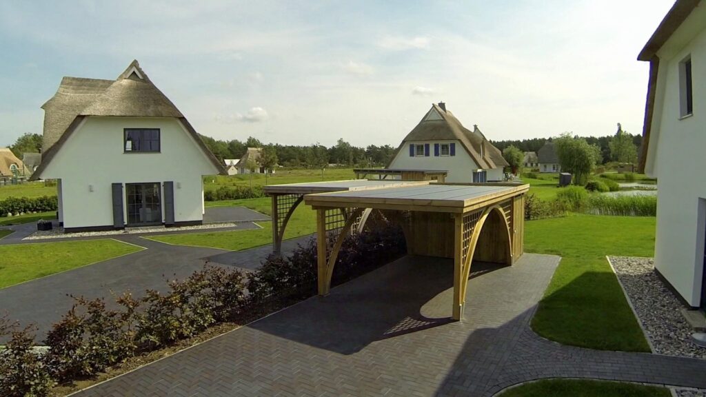 Rundbogen-Carport aus Holz für ein Fahrzeug vor einem modernen Haus mit Reetdach